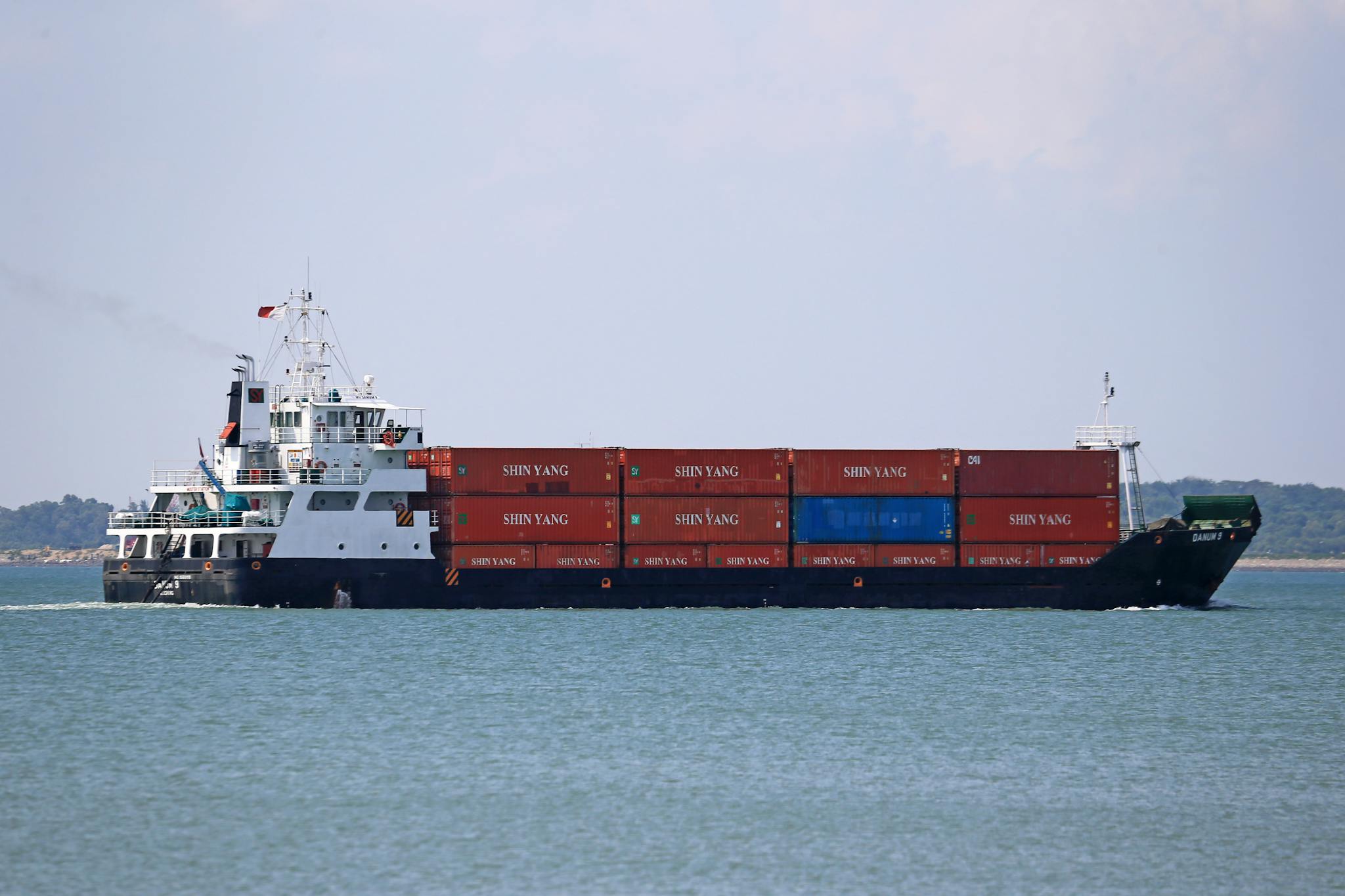 A cargo ship carrying shipping containers sails across the ocean under bright daylight.
