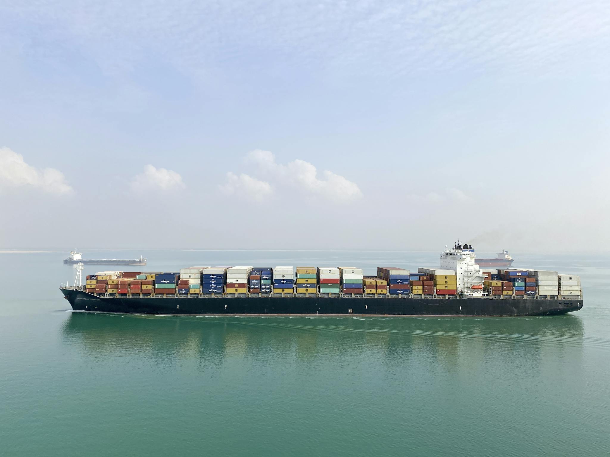 Aerial view of a large cargo ship filled with containers sailing on a calm sea under a clear sky.
